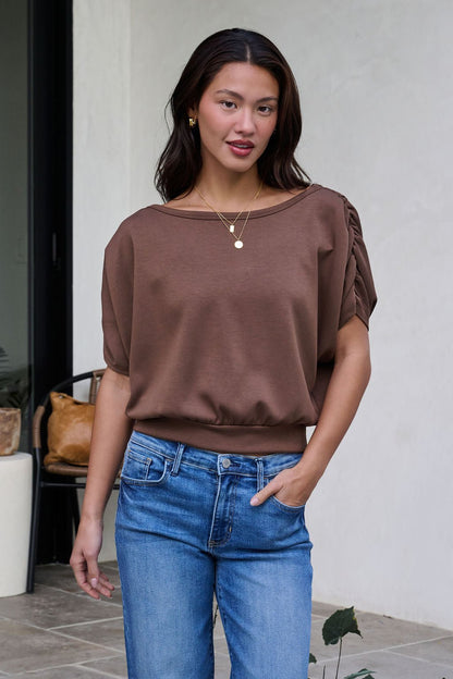 A woman smiles outdoors with one hand in her pocket, wearing a Brown Shirred Sleeve Boatneck Top and blue jeans near a chair.