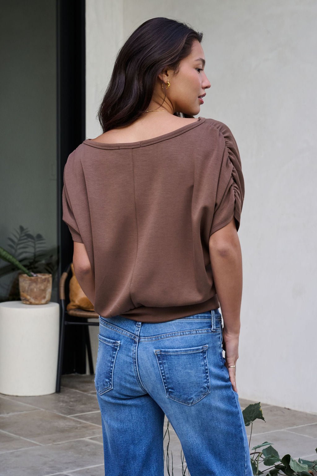 A woman stands outdoors facing away, wearing the Brown Shirred Sleeve Boatneck Top and blue jeans. Plants and a white wall are behind her.