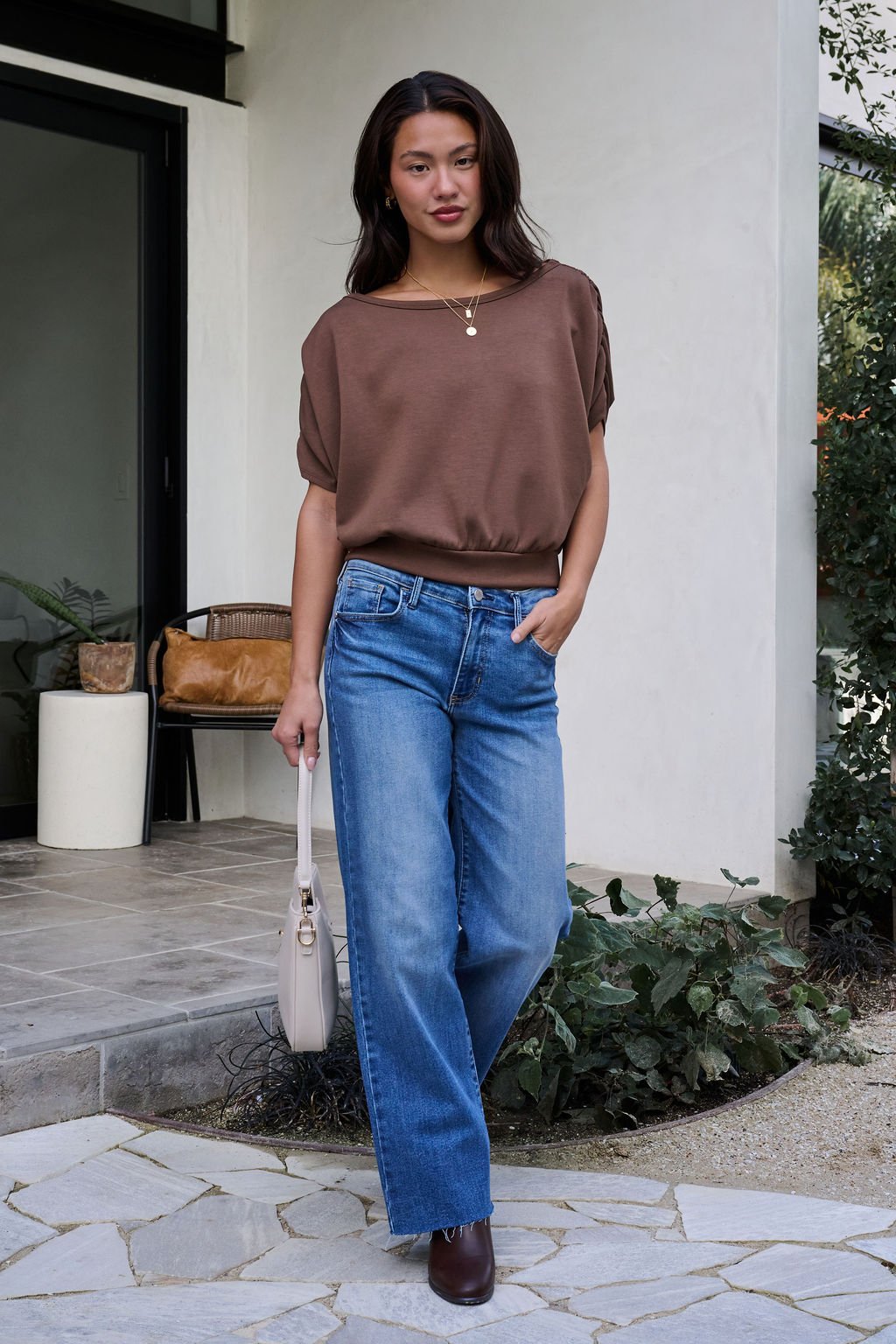 A woman in a Brown Shirred Sleeve Boatneck Top and blue jeans stands outside, holding a beige handbag and wearing brown shoes.