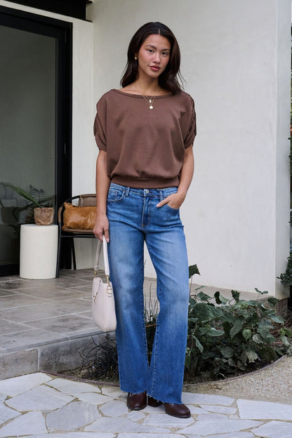 A woman stands outside a modern building wearing a Brown Shirred Sleeve Boatneck Top, blue jeans, brown shoes, and a light handbag.