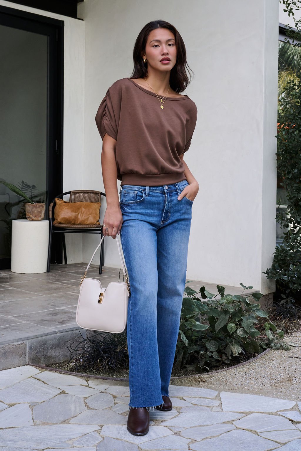 A woman stands outside a house in a Brown Shirred Sleeve Boatneck Top, blue jeans, brown shoes, and holding a white handbag.