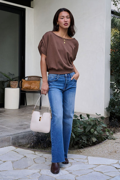 A woman stands outside a house in a Brown Shirred Sleeve Boatneck Top, blue jeans, brown shoes, and holding a white handbag.