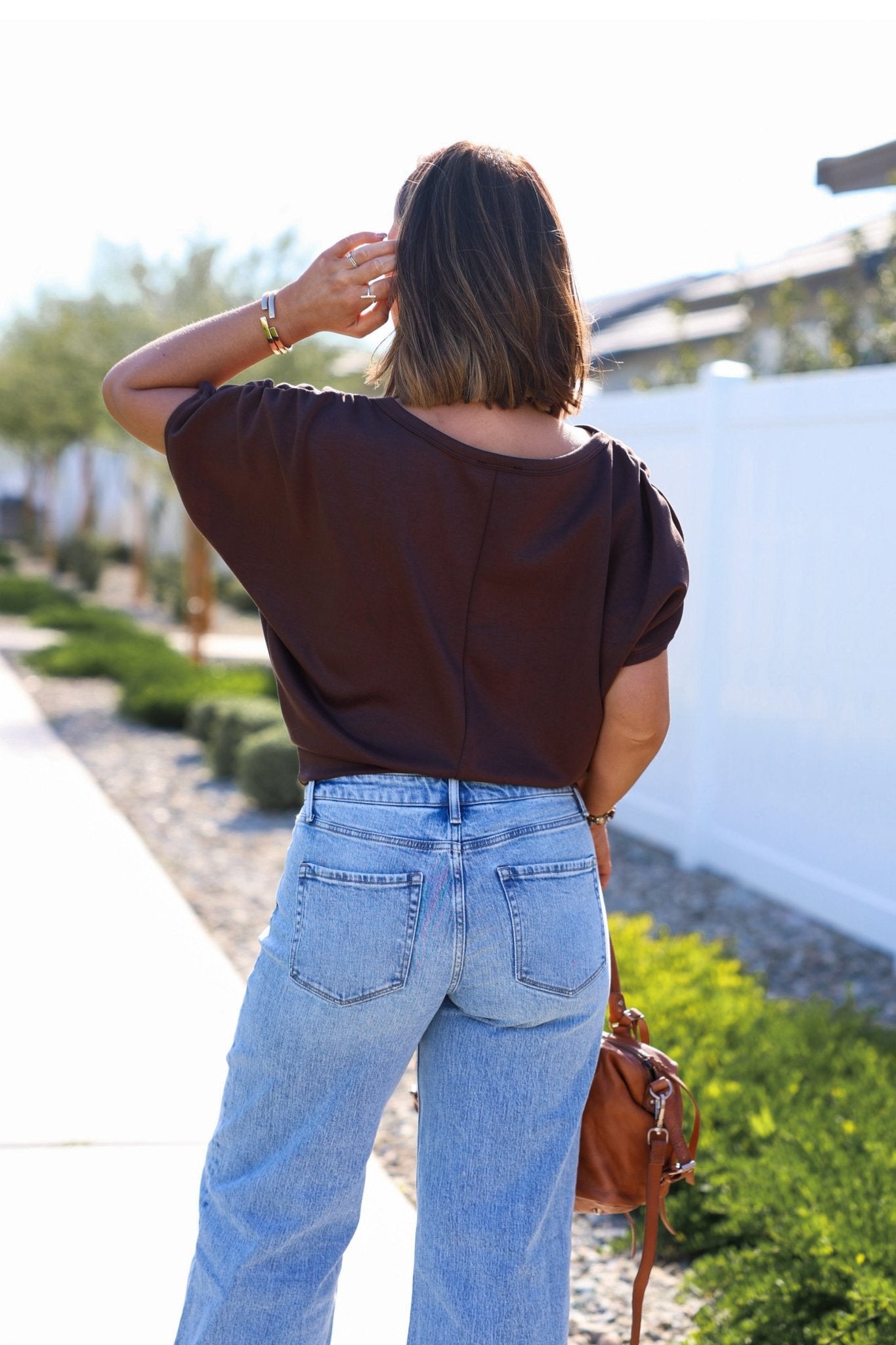 Woman with shoulder-length brown hair wears a Brown Shirred Sleeve Boatneck Top and blue jeans, standing outside on a sunny day.