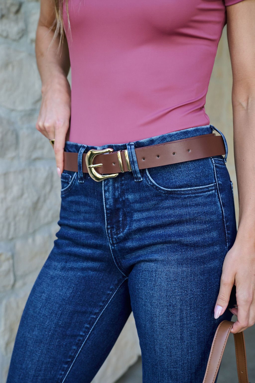 Woman in a pink top, blue jeans, and a Brown Vegan Faux Leather Belt with gold buckle, by a stone wall.