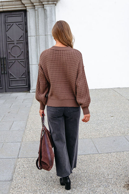 A woman in a Brown Waffle Knit Sweater and black jeans walks on pavement carrying a large brown bag.