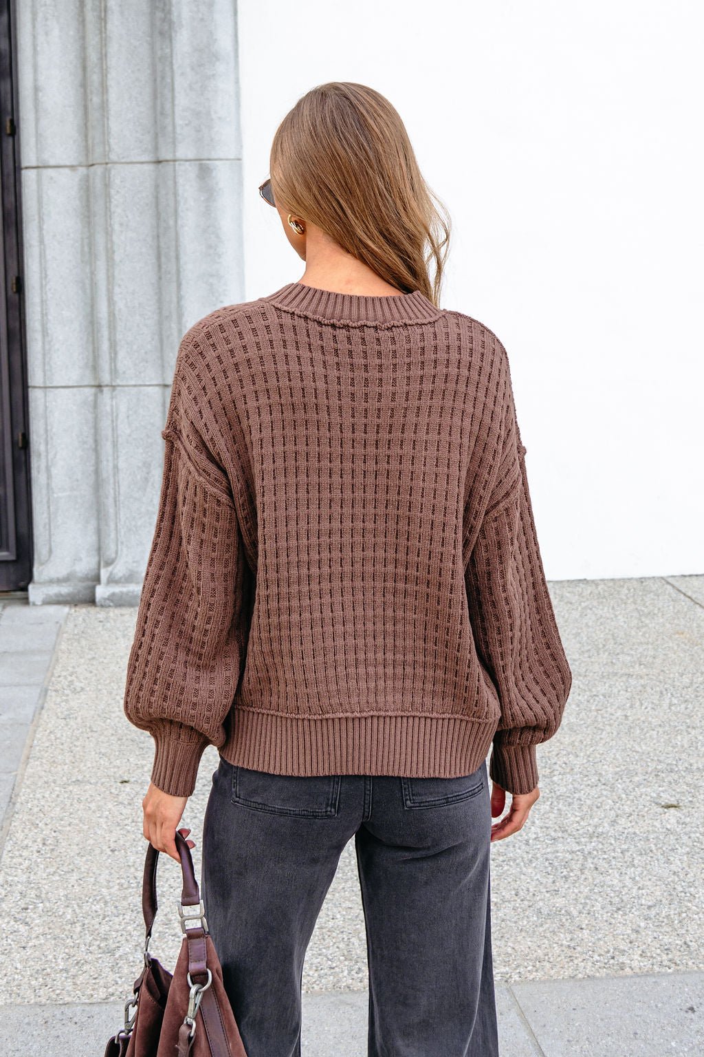 A person with long hair wears a Brown Waffle Knit Sweater and dark pants, holding a bag, standing outdoors facing away.