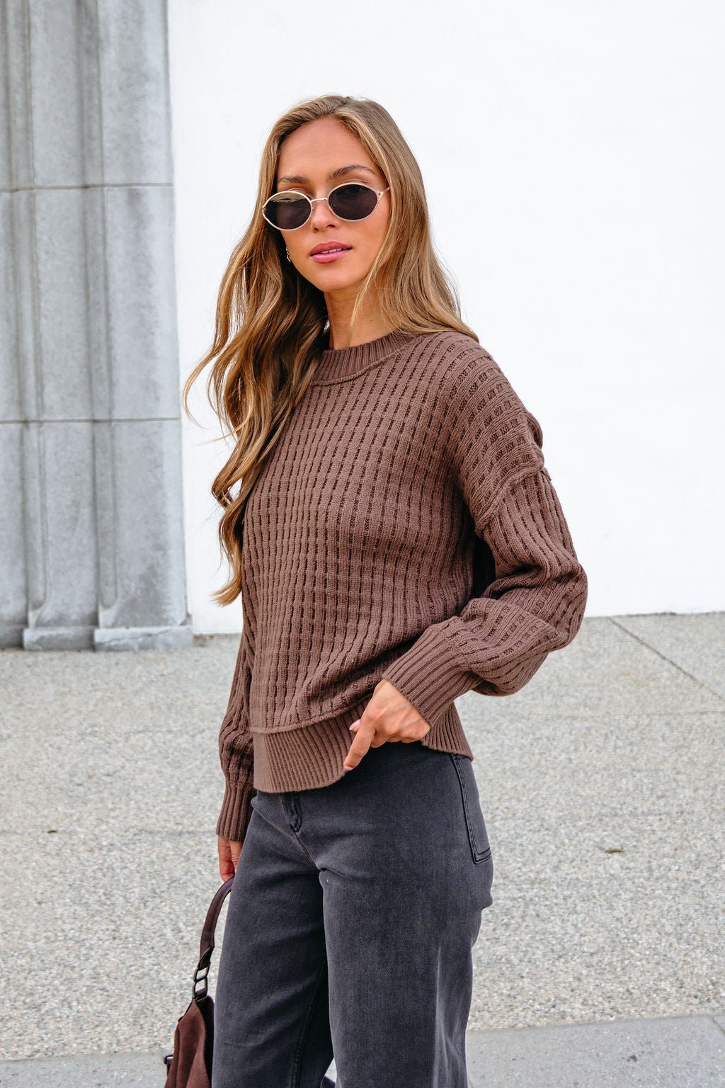 Woman with long hair in sunglasses wears a Brown Waffle Knit Sweater while standing outside by a gray stone wall.