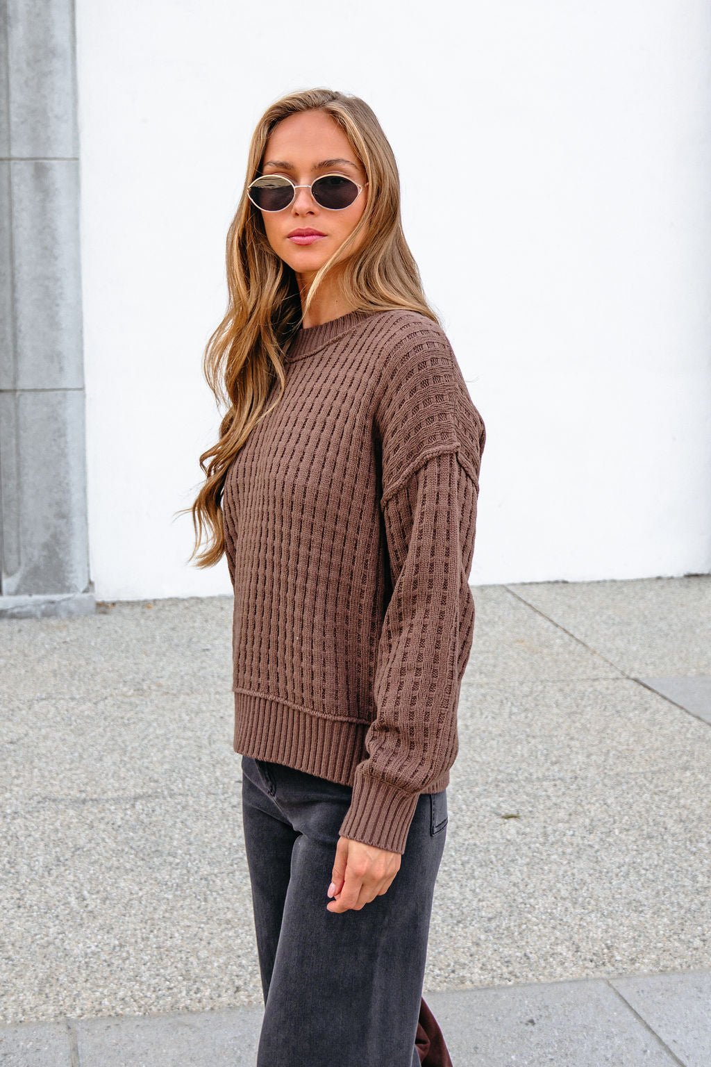 Woman with long hair in sunglasses wears a Brown Waffle Knit Sweater and dark pants outside against a white wall.