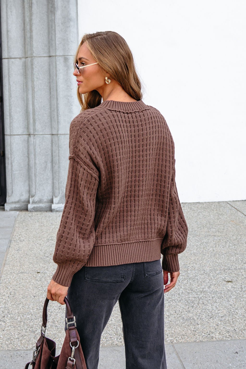 Woman with long brown hair in sunglasses and jeans, holding a bag, wears our Brown Waffle Knit Sweater outdoors.