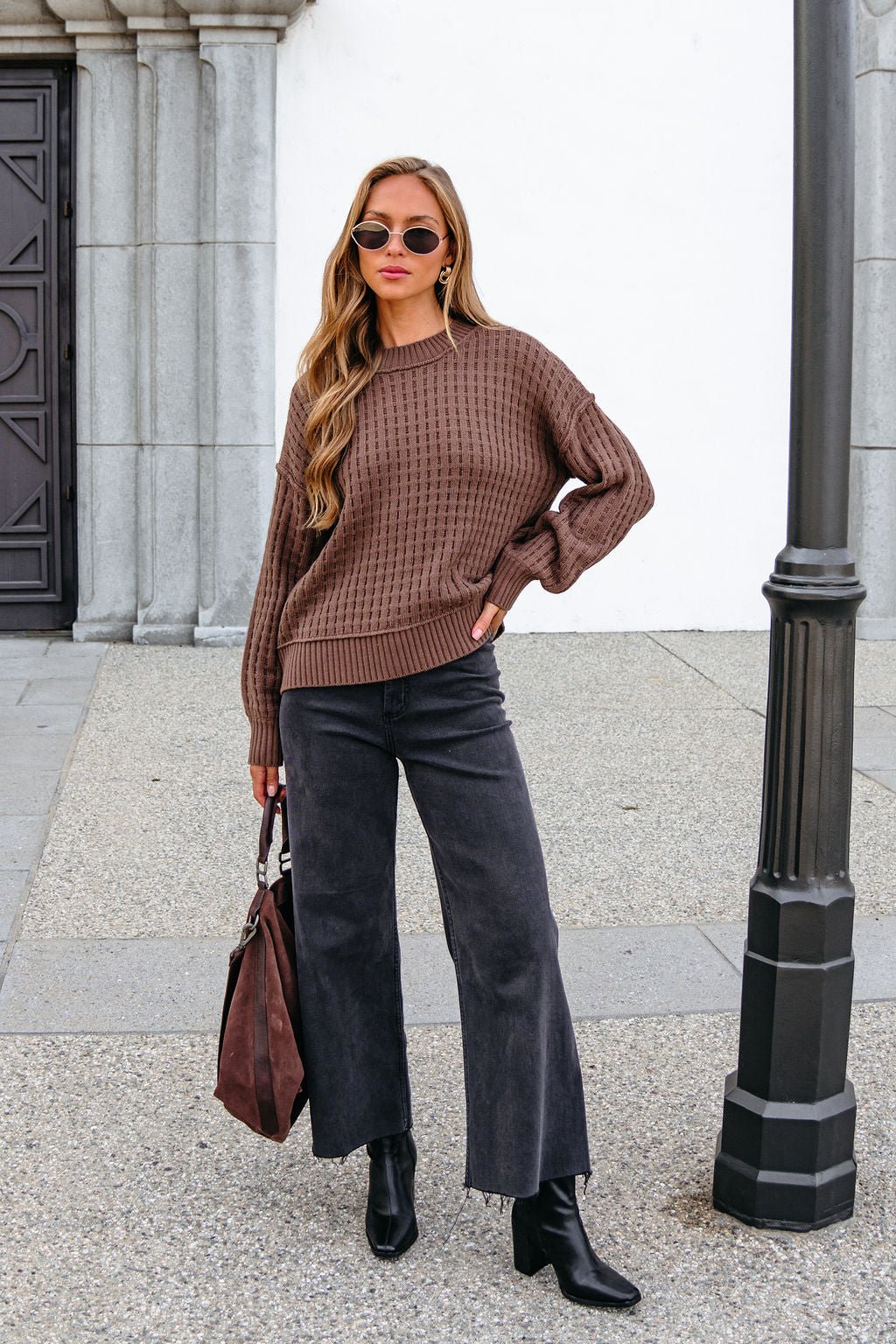 Woman in a Brown Waffle Knit Sweater, black jeans, and boots holds a brown bag by a lamppost, radiating effortless cool.