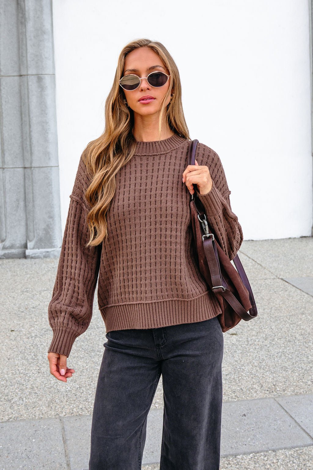 Woman in a Brown Waffle Knit Sweater, sunglasses, black pants, and brown bag stands outside by a building.