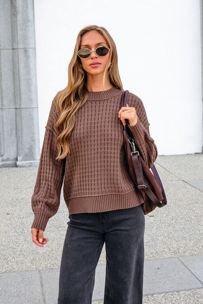 Woman in a Brown Waffle Knit Sweater, sunglasses, black pants, and brown bag stands outside by a building.