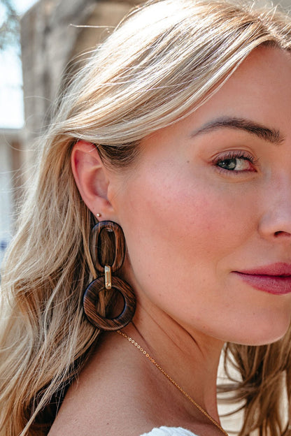 A woman with blonde hair models Brown Wooden Linked Dangle Earrings and a gold necklace in natural sunlight, facing the camera.