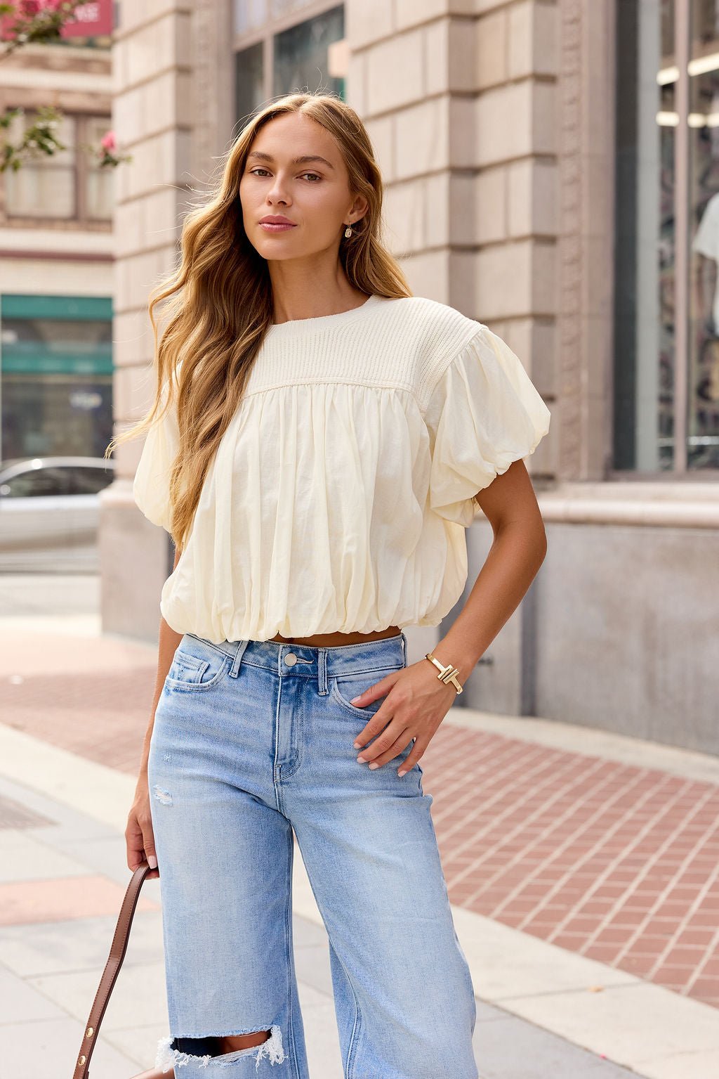 Woman with long hair wears the Bubble Hem Short Sleeve Top in Ivory and ripped jeans, holding a brown bag on the city sidewalk.