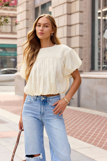 Woman with long hair wears the Bubble Hem Short Sleeve Top in Ivory and ripped jeans, holding a brown bag on the city sidewalk.