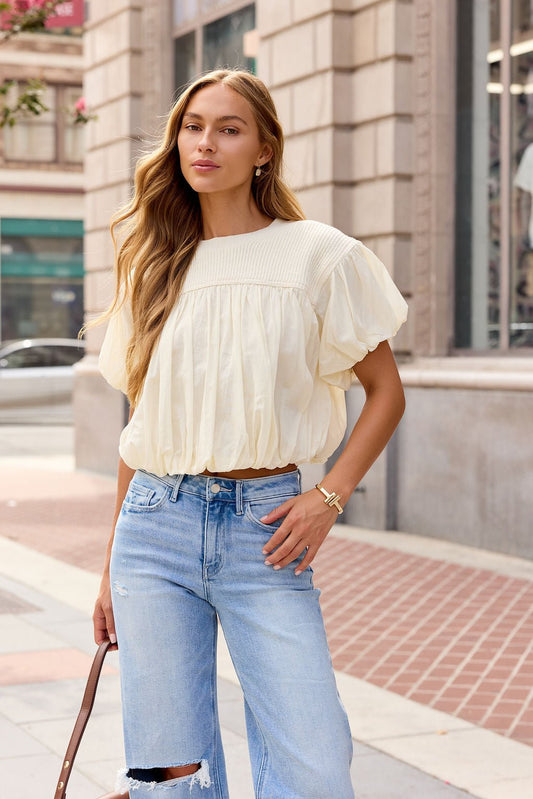 Woman in the Bubble Hem Short Sleeve Top - Ivory stands on a city sidewalk, holding a brown strap and wearing ripped jeans.