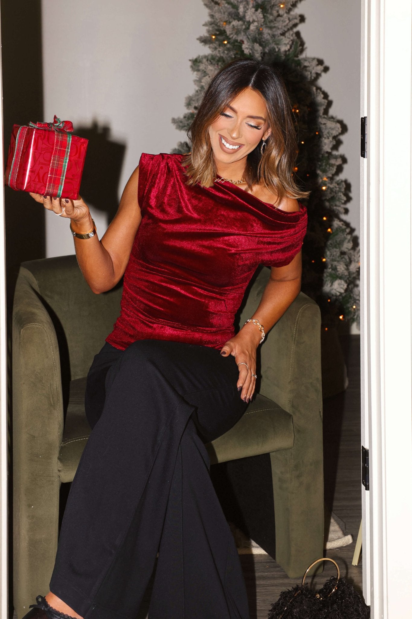 A woman in a Burgundy Asymmetrical Ruched Velvet Top and black pants sits on a green chair, smiling with a wrapped gift by the Christmas tree.