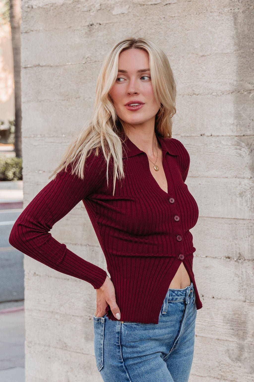 A woman with long blonde hair models the Burgundy Button Down Ribbed Top with blue jeans against a concrete wall.