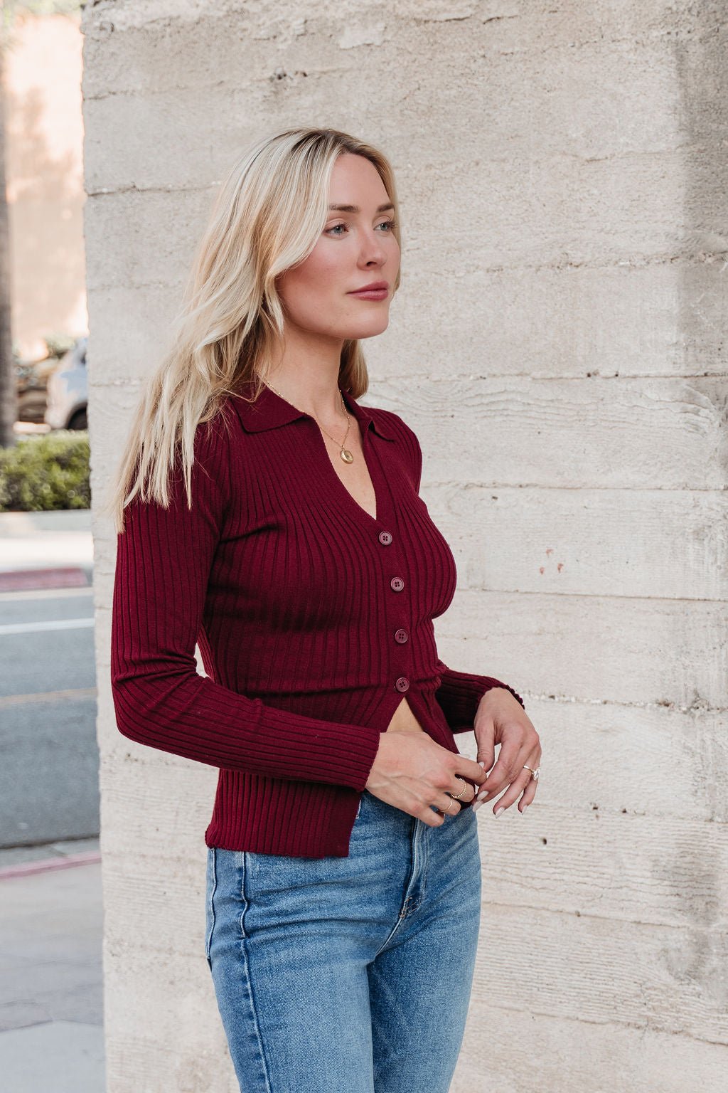 A woman in a Burgundy Button Down Ribbed Top and blue jeans stands outside, showcasing cozy fall style.