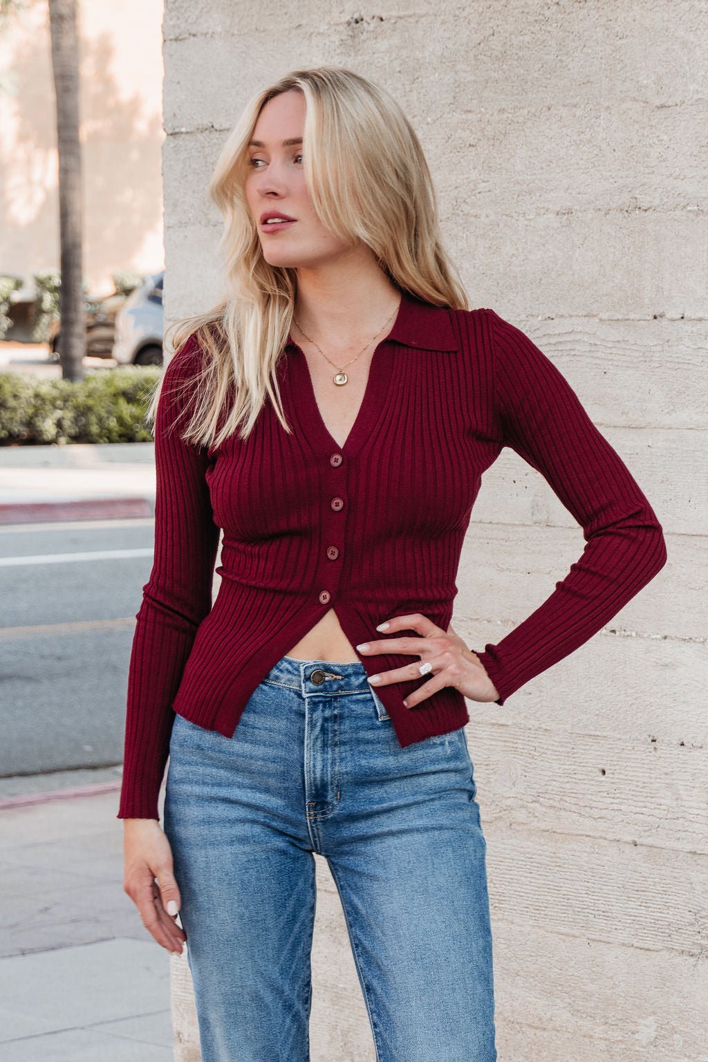 A woman in the Burgundy Button Down Ribbed Top | Pre Order and blue jeans showcases fall style by a concrete wall.