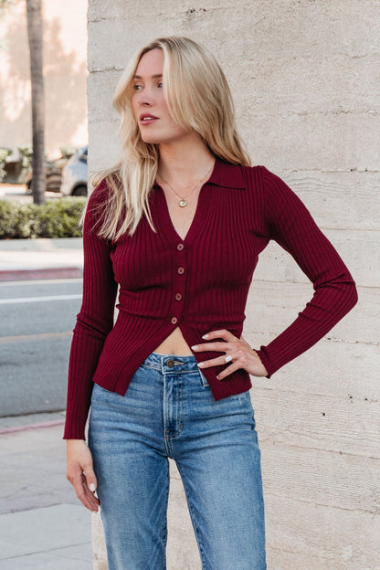 A woman in the Burgundy Button Down Ribbed Top | Pre Order and blue jeans showcases fall style by a concrete wall.