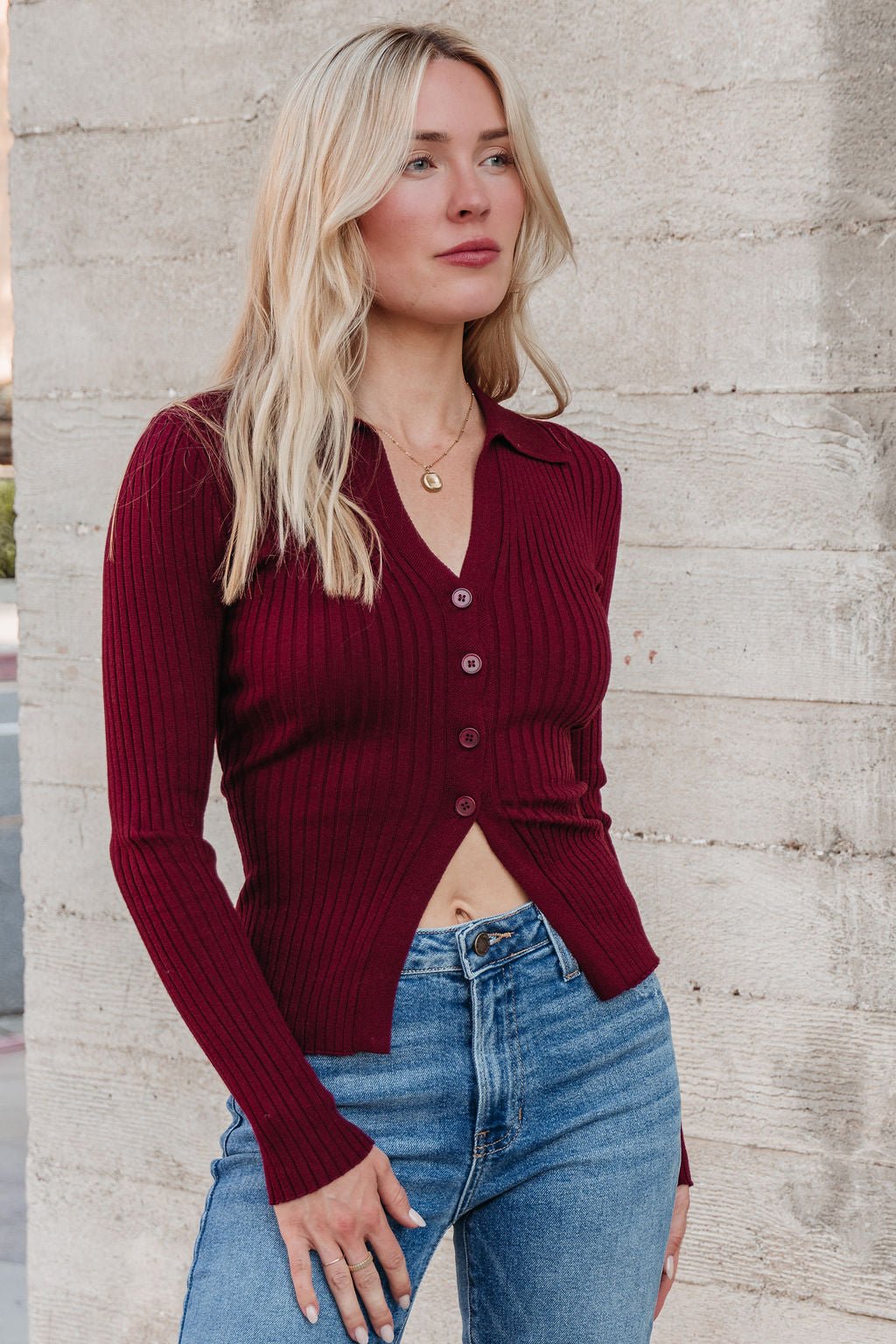 A woman with long blonde hair wears the Burgundy Button Down Ribbed Top and blue jeans, posing by a concrete wall for fall style.
