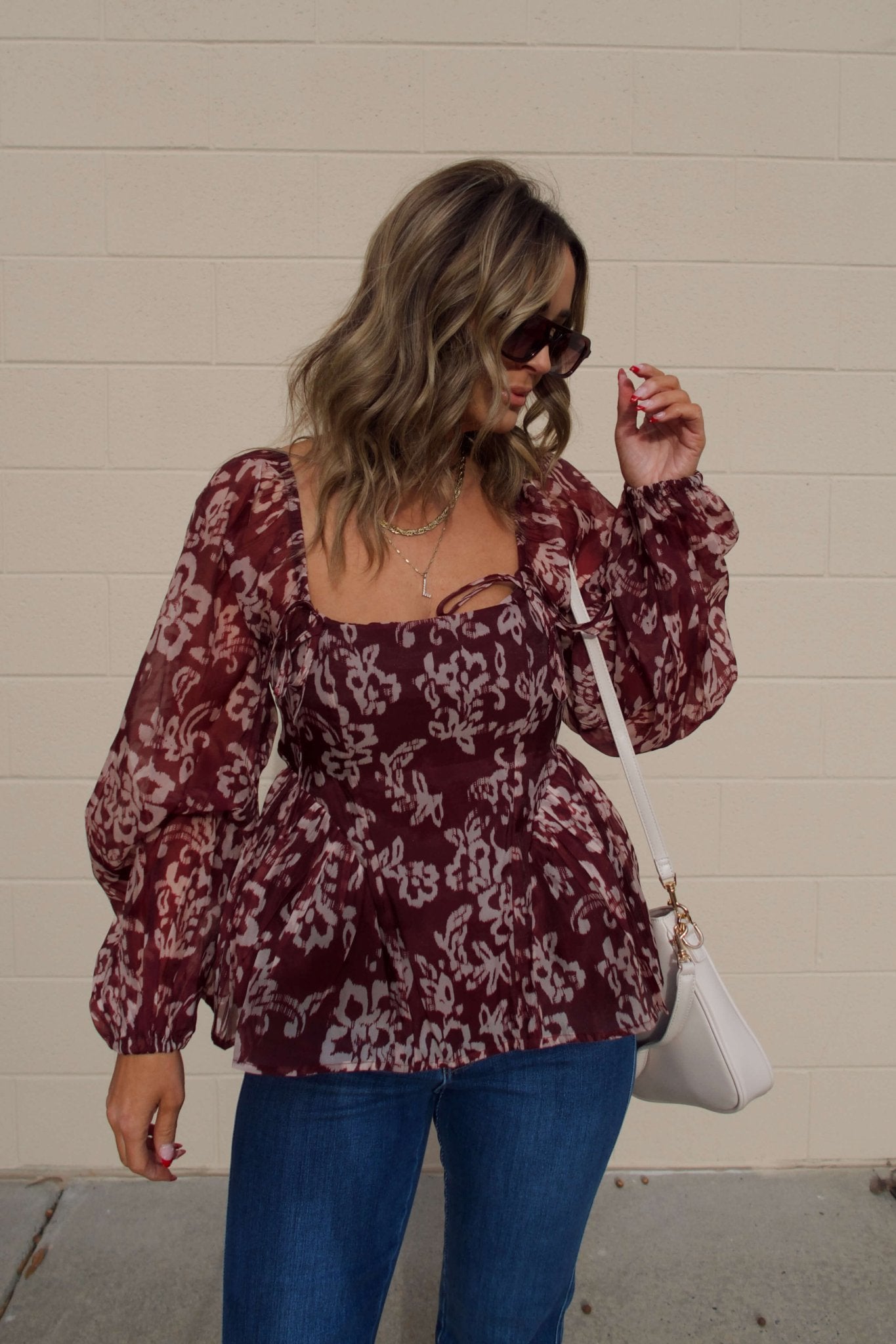 Woman with wavy hair in sunglasses wears a Burgundy Floral Chiffon Peplum Top and blue jeans, carrying a white purse.