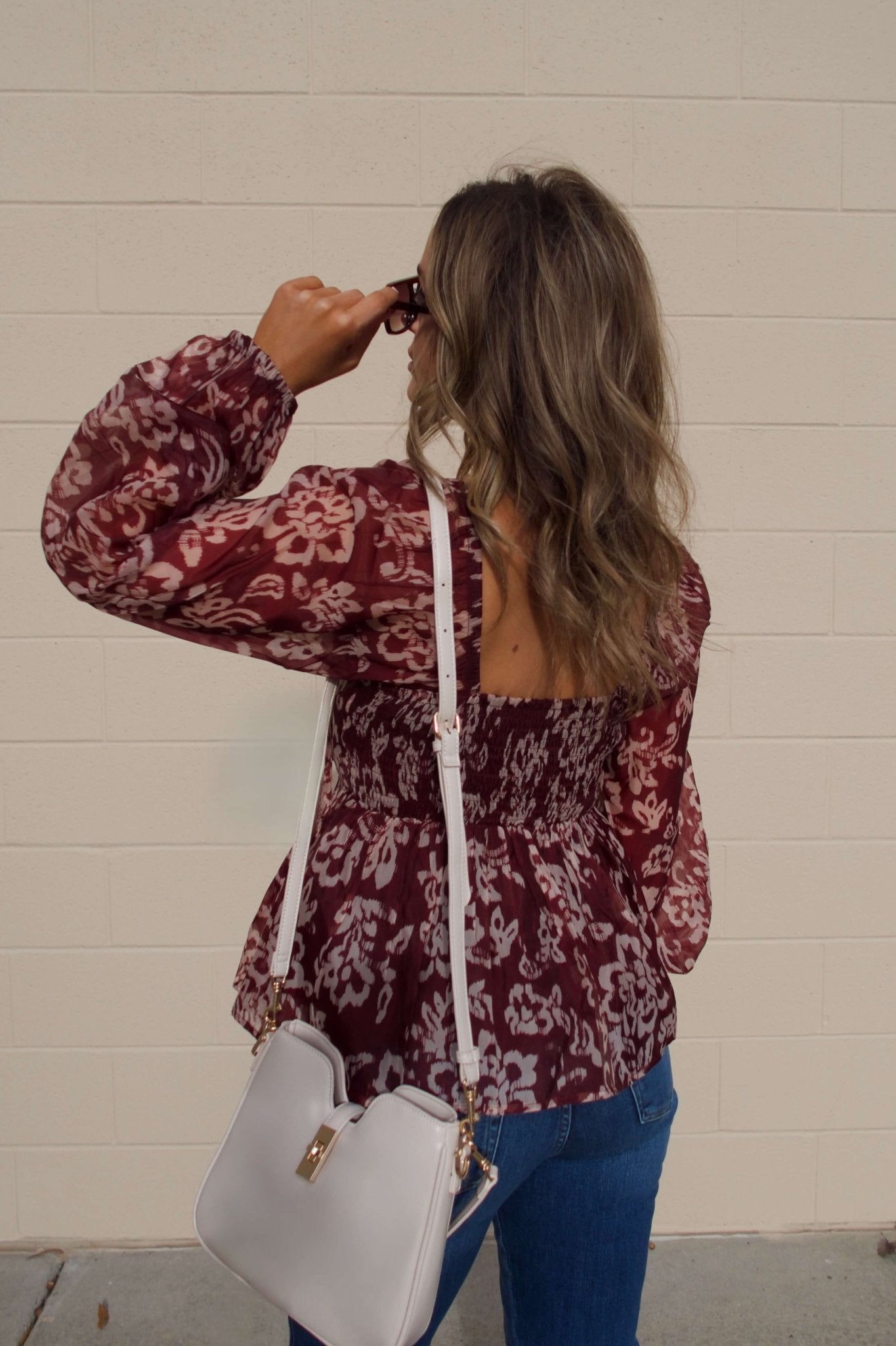 A woman in sunglasses and a Burgundy Floral Chiffon Peplum Top with jeans and a white purse stands facing a beige wall.