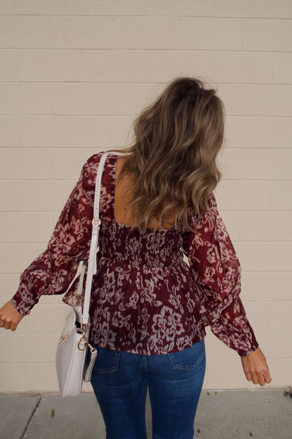 Woman with wavy hair in a Burgundy Floral Chiffon Peplum Top, jeans, and white bag stands facing a beige wall, seen from behind.