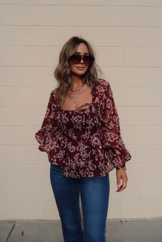 Woman in sunglasses smiles slightly, wearing a Burgundy Floral Chiffon Peplum Top and blue jeans against a beige brick wall.