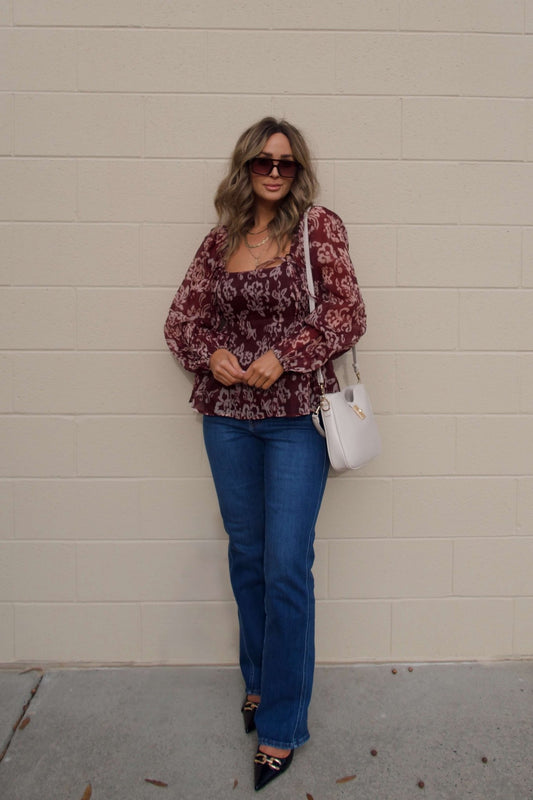 Woman in a Burgundy Floral Chiffon Peplum Top, blue jeans, and black heels with a white purse by a beige brick wall.