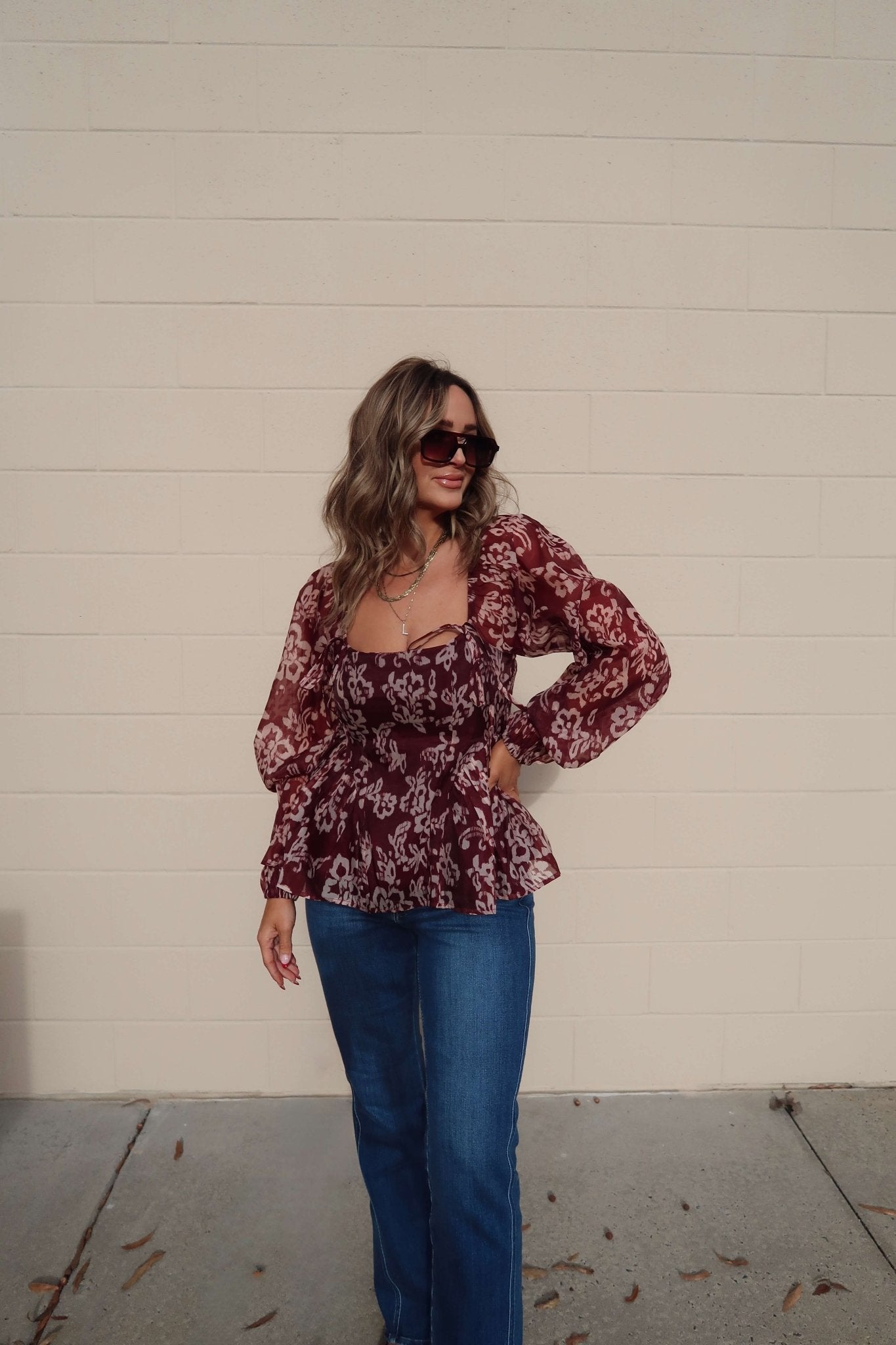 Woman smiles in a Burgundy Floral Chiffon Peplum Top and blue jeans, standing against a beige brick wall.