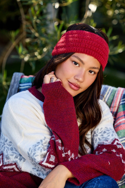 A woman in a Burgundy Fuzzy Soft Cable Knit Headband and maroon sweater sits outdoors, chin on hand, with a patterned blanket behind her.