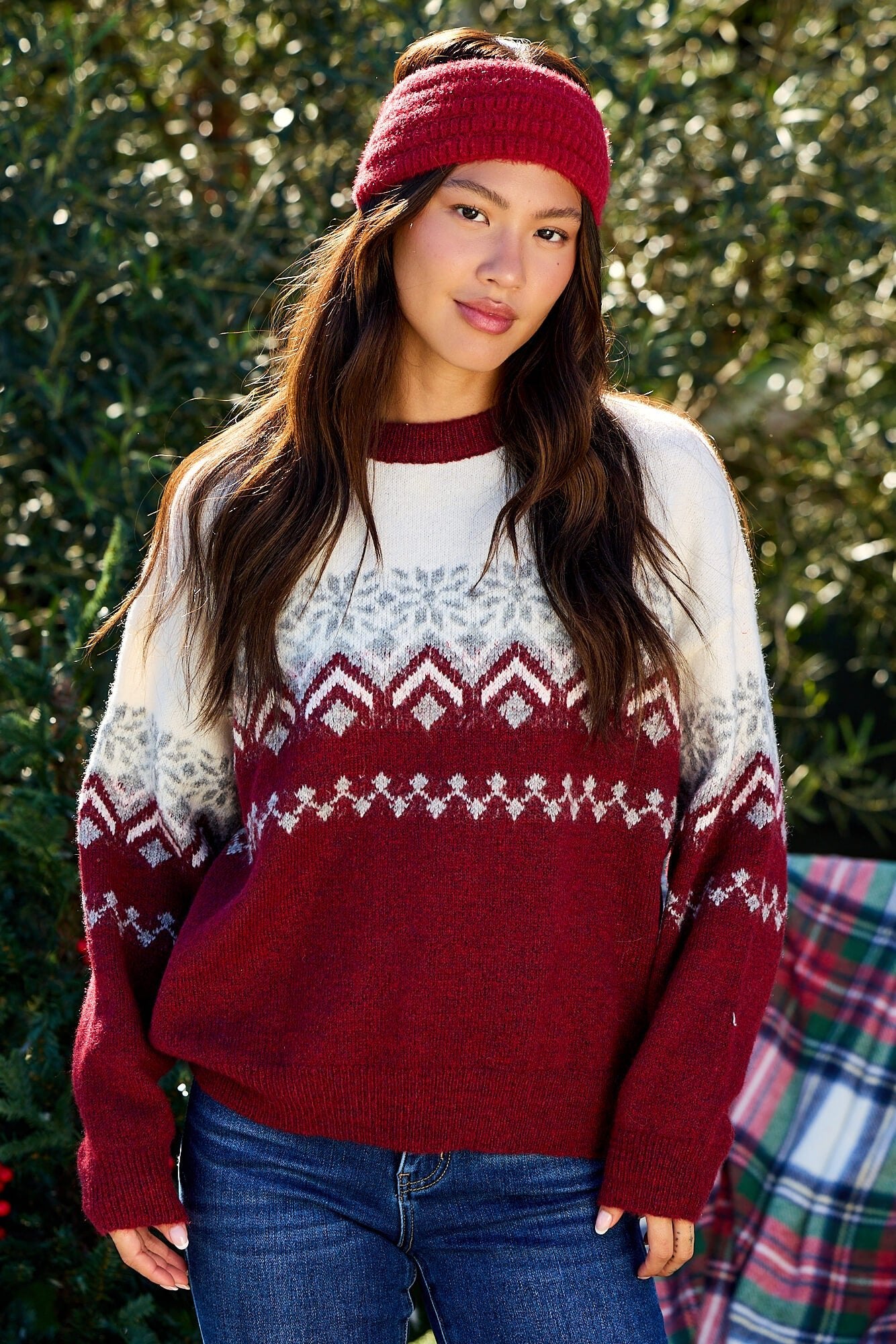Woman outdoors in a Burgundy Mix Fair Isle Sweater, blue jeans, and red headband, standing before green foliage.
