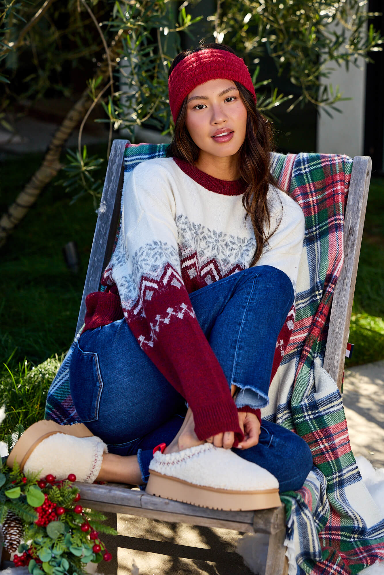 Woman in a red beanie and Burgundy Mix Fair Isle Sweater sits outdoors on a chair with a plaid blanket, near festive greenery.