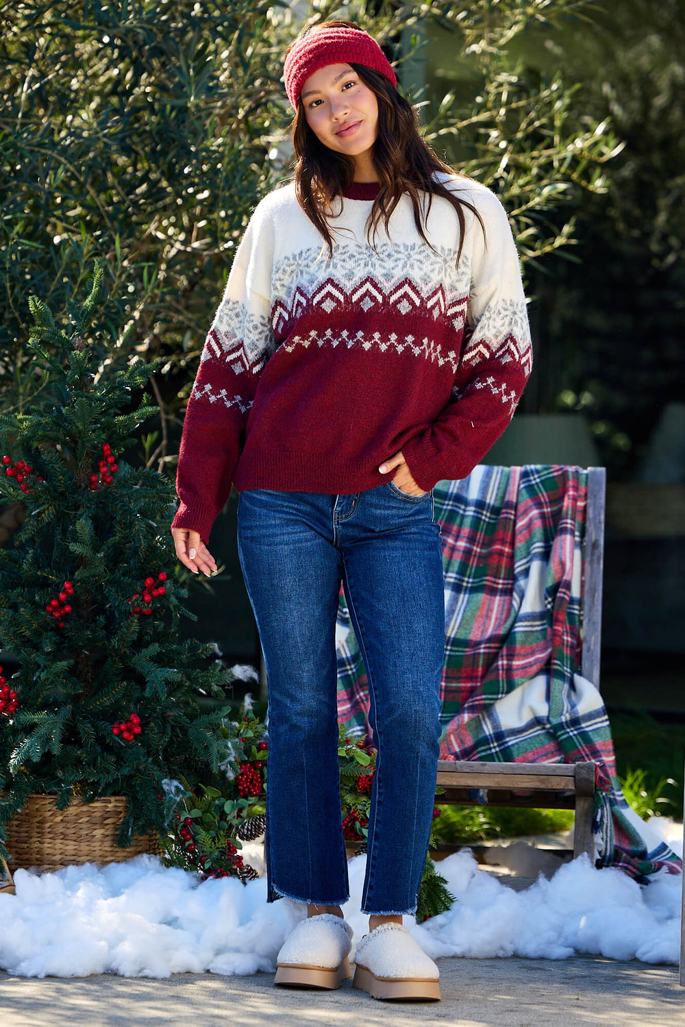 A woman in a Burgundy Mix Fair Isle Sweater, jeans, and red beanie stands by a festive holiday display with greenery and snow.