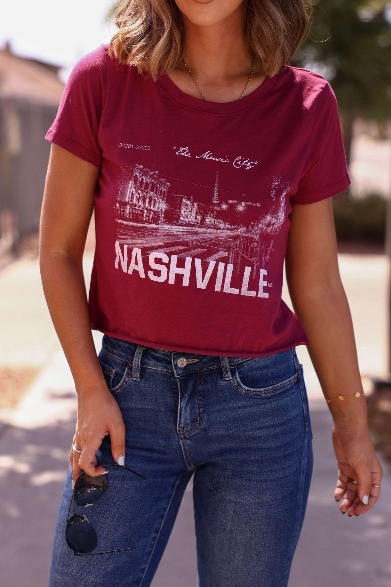 Woman in a burgundy Nashville Nights graphic tee and blue jeans holding sunglasses, standing outdoors on a sunny day.