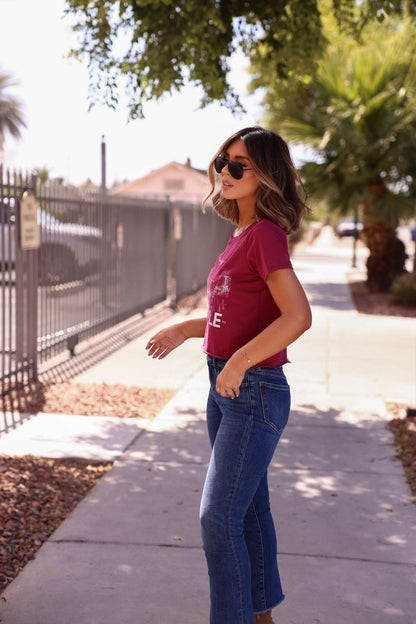 Woman in a Burgundy Nashville Nights Graphic Tee, sunglasses, and jeans stands on a sunny sidewalk with trees and a fence.