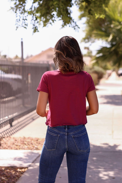 A woman with shoulder-length hair wears a Burgundy Nashville Nights Graphic Tee and blue jeans, standing on a sidewalk, back to camera.
