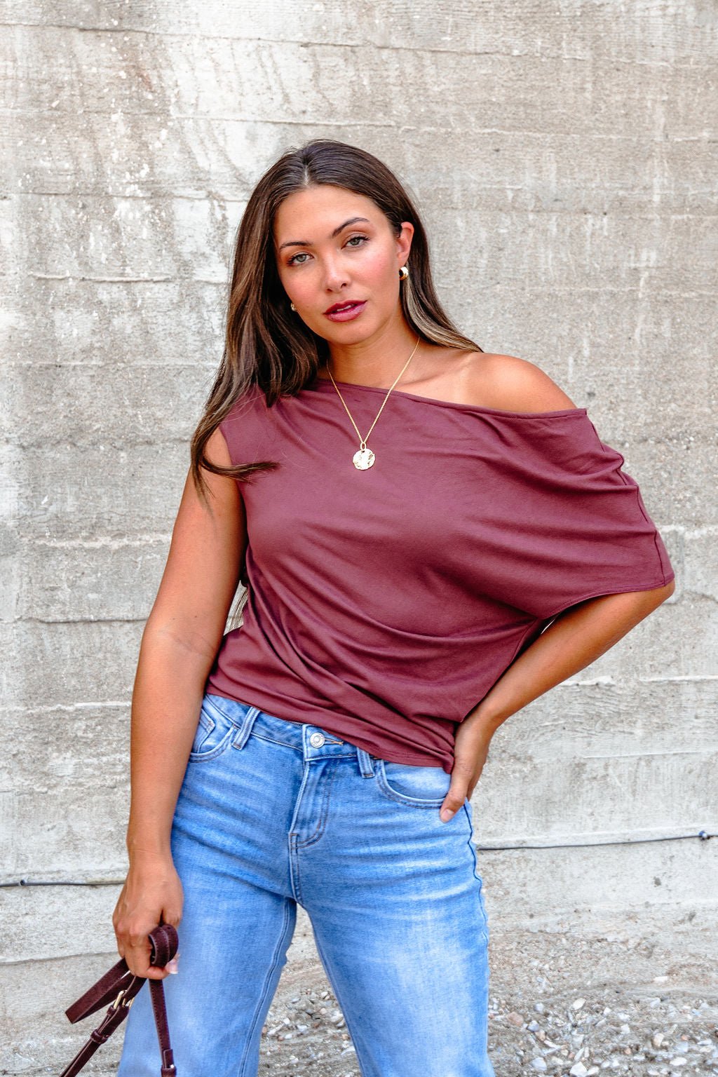 Woman with long brown hair wears a Burgundy Off The Shoulder Top - DOORBUSTER and blue jeans, standing against a concrete wall with sunglasses.