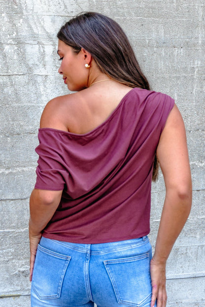 A woman in a Burgundy Off The Shoulder Top - DOORBUSTER and blue jeans stands with her back to the camera facing a concrete wall.
