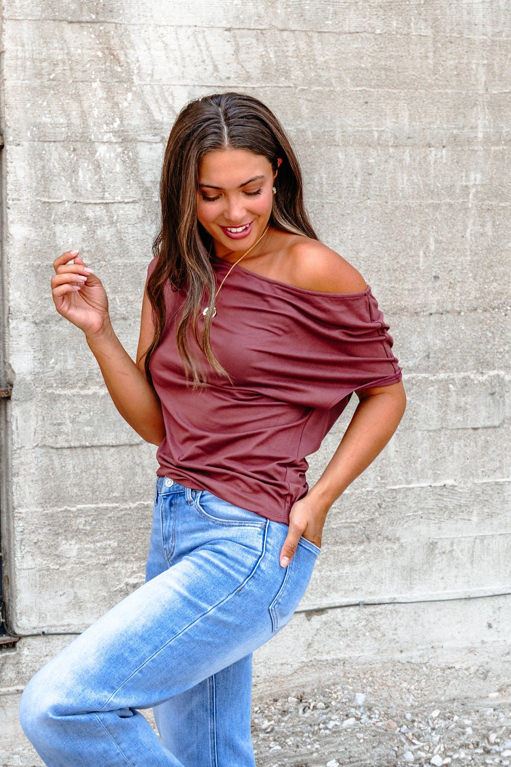 Woman in a Burgundy Off The Shoulder Top - DOORBUSTER and blue jeans stands against a concrete wall, smiling with one hand in pocket.
