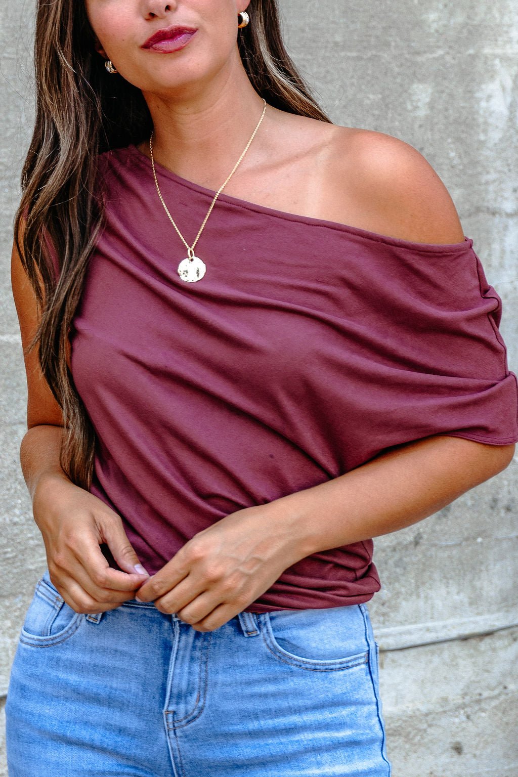 Woman in a burgundy off the shoulder top and light blue jeans, wearing a gold pendant necklace, against a gray background.