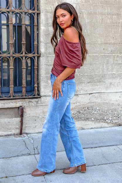 Woman with long brown hair in a Burgundy Off The Shoulder Top, blue jeans, and brown heels stands by a concrete wall and window.
