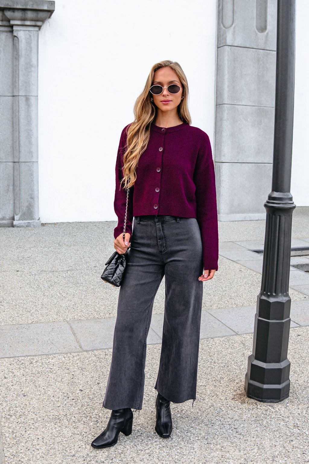 Woman in a Burgundy Ribbed Button Down Cardigan, black jeans, boots, and sunglasses stands by a lamppost on a city sidewalk.