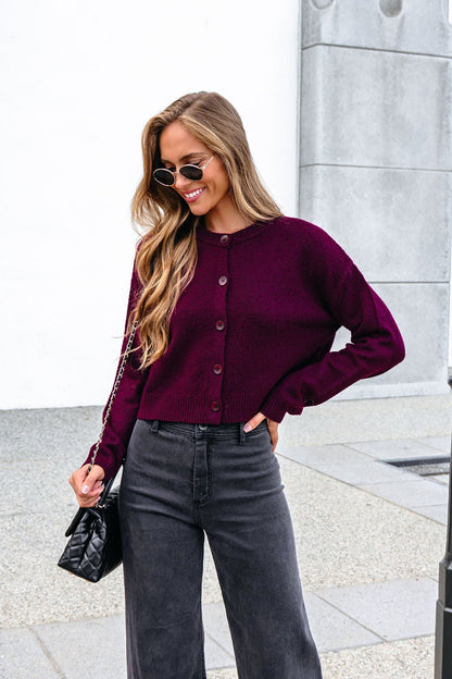 Smiling woman in sunglasses wears a Burgundy Ribbed Button Down Cardigan, holding a black purse by a white outdoor wall.