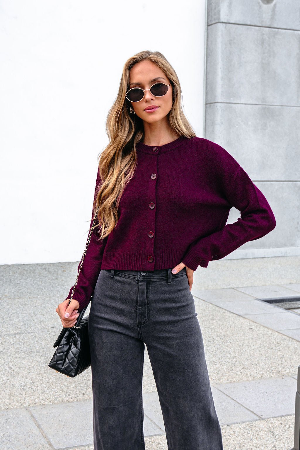 Woman in sunglasses wears a Burgundy Ribbed Button Down Cardigan and black pants, holding a black quilted purse outdoors.