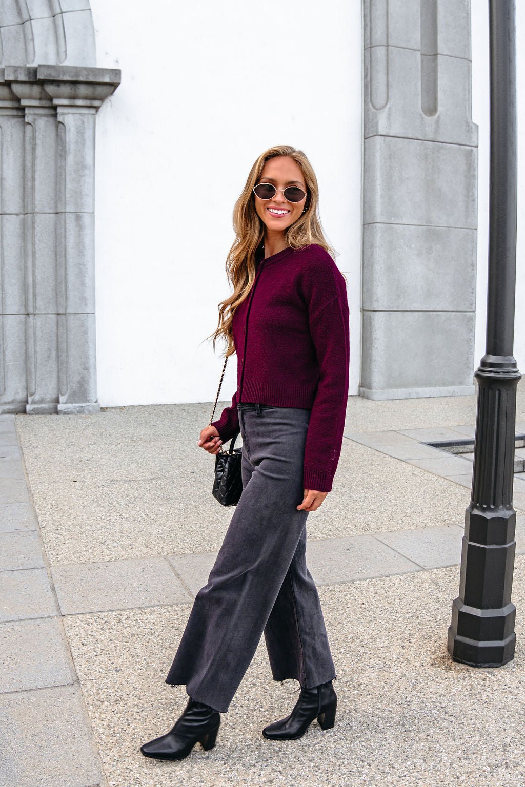 Woman in sunglasses wears a Burgundy Ribbed Button Down Cardigan, gray pants, and black boots, holding a black purse outside a gray building.