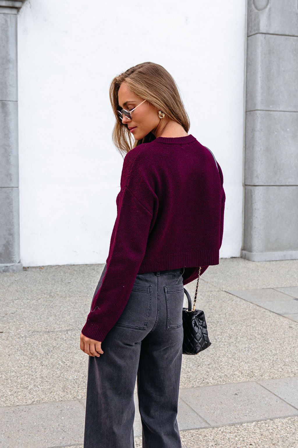 Woman in sunglasses, Burgundy Ribbed Button Down Cardigan, stands outdoors with a quilted purse and gray high-waisted pants.
