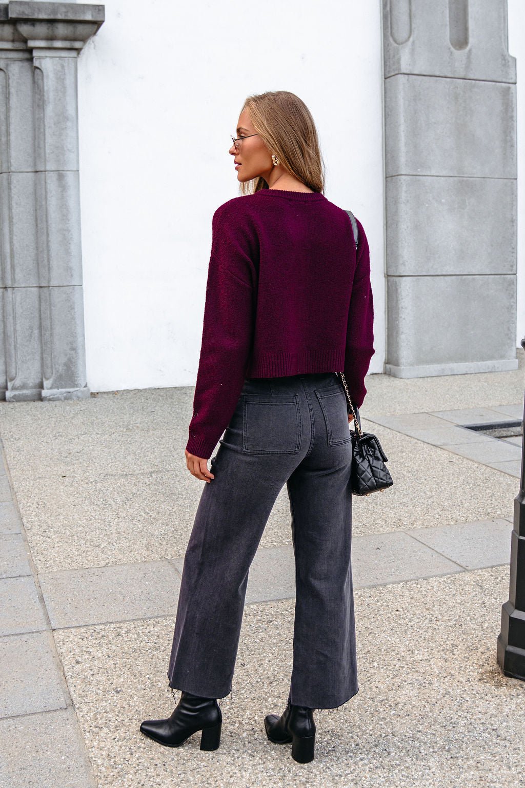 Woman with long hair wears Burgundy Ribbed Button Down Cardigan, dark jeans & boots, standing outdoors facing away from the camera.